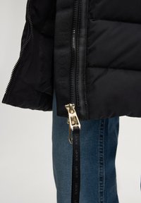 Zwart donsjack met een glanzende ritssluiting, voorzien van een gestructureerde voering en opvallende merkdetails langs de rits. Daaronder zijn blauwe spijkerbroeken zichtbaar.