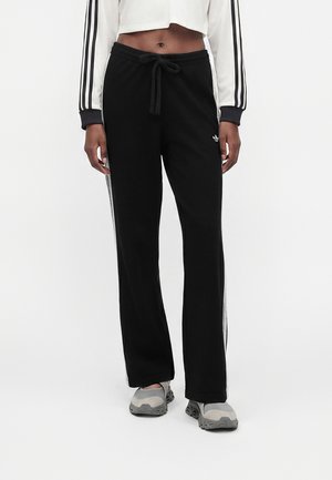 Persoon draagt zwarte Adidas trainingsbroek met witte strepen, taille met trekkoord, witte cropped top met zwarte strepen op de mouwen en grijze sportschoenen.