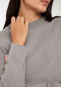 Szary sweter z prążkowanym dekoltem i mankietami. Materiał wygląda na miękki i ma gładką fakturę. Widoczna ręka z zadbanymi paznokciami dotyka swetra.