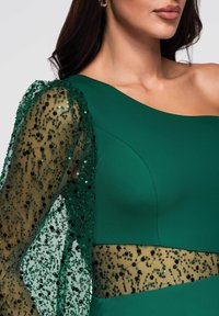 Robe verte avec un corsage structuré et une manche longue unique en tissu transparent ornée de sequins noirs et de motifs complexes.