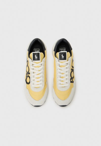 Dzelteni apavi ar baltām auklām un melniem akcentiem, uz kuriem redzams izteikts logo. Iestatīti uz balta fona.