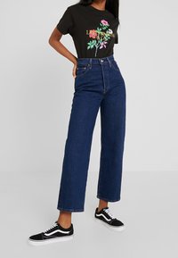 Czarny graficzny t-shirt z kwiatowym wzorem i żółtym napisem, połączony z wysokim stanem, szerokimi granatowymi dżinsami i czarnymi płóciennymi trampkami.