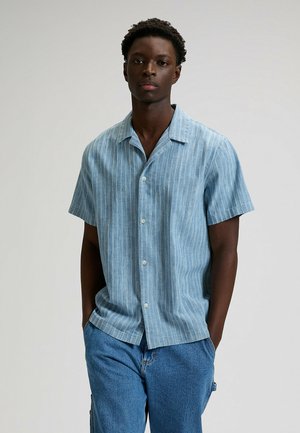 Hombre joven que lleva una camisa azul claro de manga corta con botones y rayas verticales, y jeans azules, de pie con las manos en los bolsillos contra un fondo liso.