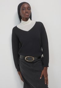 Maglione nero a v-neck con texture a coste, sovrapposto a un dolcevita grigio chiaro. Gonna di jeans scuri e una cintura nera lucida con una fibbia ovale dorata.