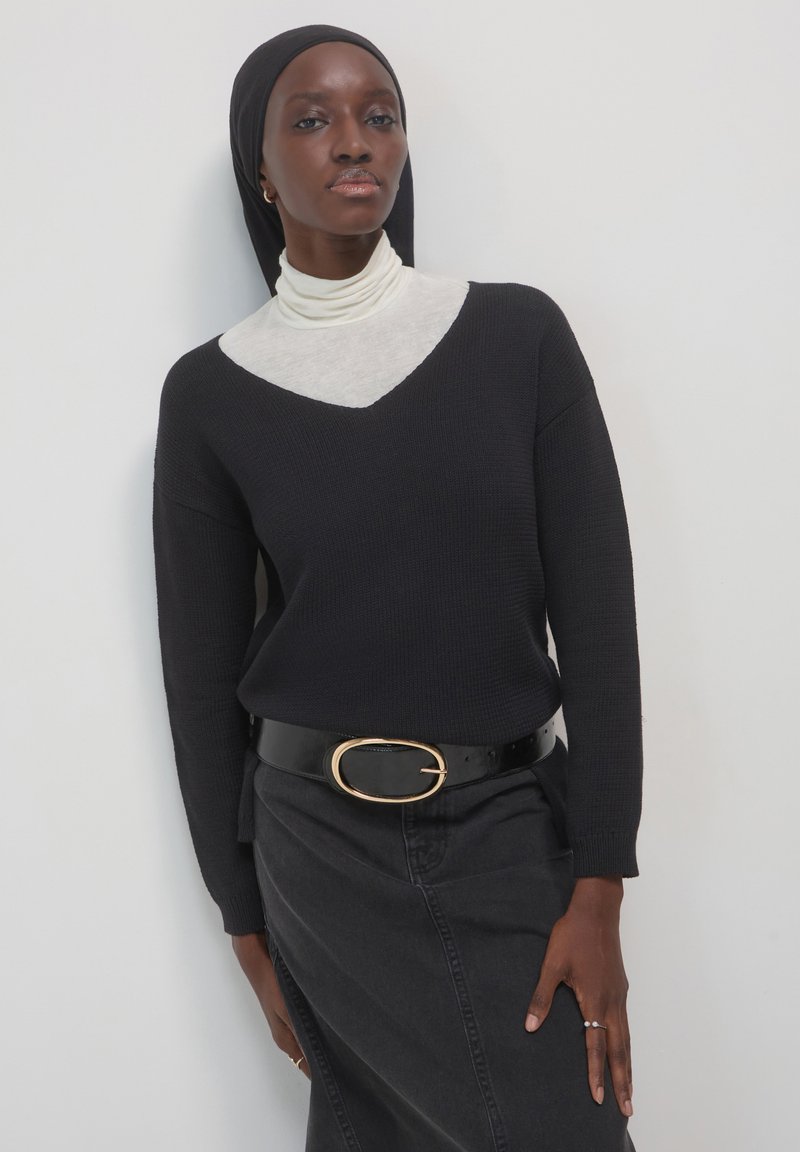 Maglione nero a v-neck con texture a coste, sovrapposto a un dolcevita grigio chiaro. Gonna di jeans scuri e una cintura nera lucida con una fibbia ovale dorata.