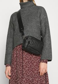 Bolso bandolera de cuero negro con cremalleras; presenta una superficie texturizada, correa ajustable y forma rectangular compacta.