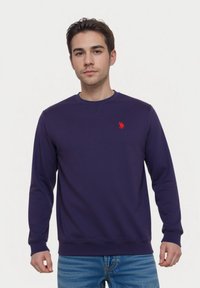Jonge man in een effen donkerblauwe sweatshirt met een klein rood geborduurd logo op de borst en blauwe jeans, staande tegen een witte achtergrond.