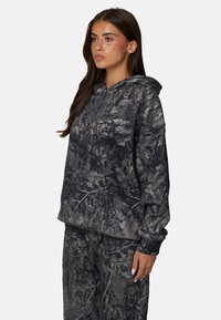 Bluza z kapturem w ciemnoszarym kolorze z wzorem w kamuflaż. Wykonana z miękkiego materiału, z kieszenią typu kangur i ściągaczami przy mankietach. Luźny krój.