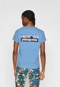 Μπλε βαμβακερό T-shirt με μαύρο γραφικό σχέδιο που απεικονίζει βουνά και κείμενο "patagonia", φορεμένο με πολύχρωμα σορτς με λουλουδάτο μοτίβο.