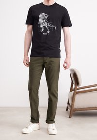 Man som bär en svart grafisk T-shirt med en mekanisk dinosaurie och ordet "BOSS", olivgröna byxor och vita sneakers, står bredvid en beige fåt ölj.