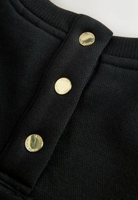 Tessuto di felpa nero con finitura strutturata, caratterizzato da tre bottoni a pressione in metallo dorato al collo per la chiusura. Dettaglio del colletto a coste.