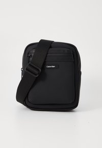 Borsa a tracolla nera in nylon con texture liscia, tasche con cerniera e tracolla regolabile. Presenta il logo Calvin Klein sul davanti.