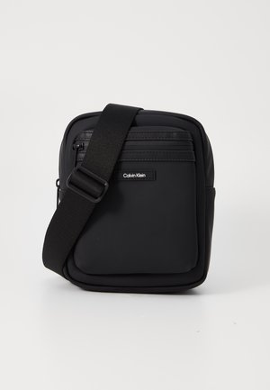 Bolso bandolera negro de nailon con textura suave, bolsillos con cremallera y correa ajustable. Presenta un logo de Calvin Klein en la parte frontal.