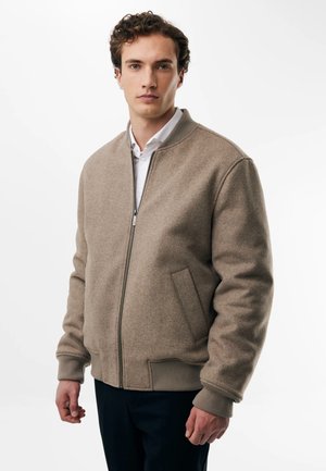 Giovane uomo con capelli corti e ricci che indossa una giacca bomber beige con zip sopra una camicia bianca, in piedi davanti a uno sfondo bianco.