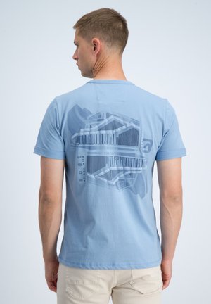 Man met kort haar van achteren gezien, draagt een lichtblauw T-shirt met een geometrisch gebouwontwerp en tekst op de achterkant, gecombineerd met beige broek.