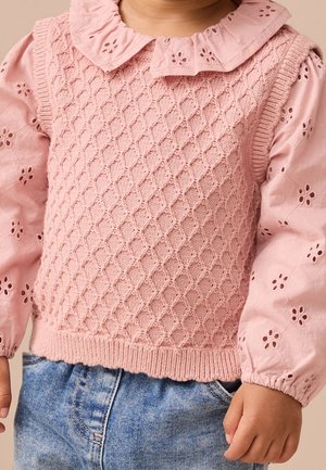 Barn iført lyserød tekstureret strikkevest over lyserød langærmet bluse med blomstrede udskæringer og blå jeans, vist fra nakke til hofter.