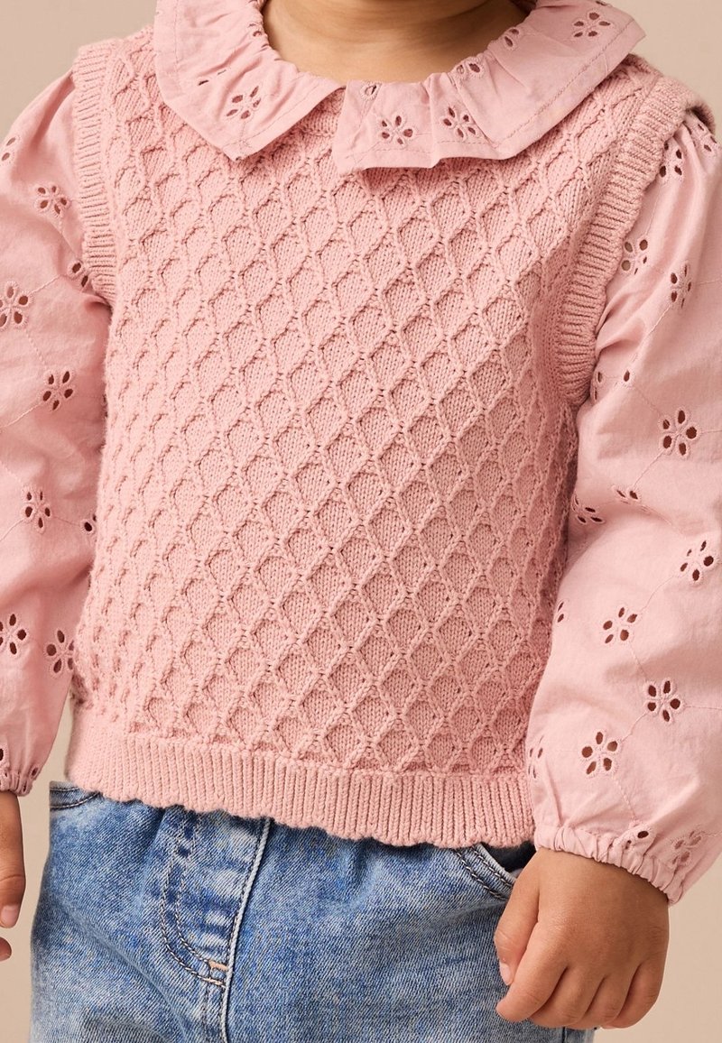 Enfant portant un gilet en tricot texturé rose sur une blouse rose à manches longues avec découpes florales et un jean bleu, présenté du cou jusqu'aux hanches.