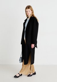 Μαύρο μακρύ παλτό με γιακα, συνδυασμένο με μπλε πουκάμισο με σχέδιο και χρυσή πλισέ φούστα. Μαύρες φλατ και μια μπλε τσάντα ολοκληρώ�νουν την εμφάνιση.