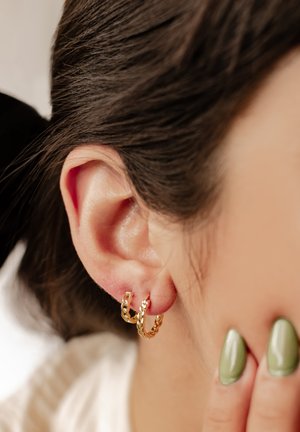 Goldfarbene Creolen mit einem gedrehten Design, die am Ohr sitzen. Kurze Haare sichtbar, ergänzt durch grünen Nagellack.