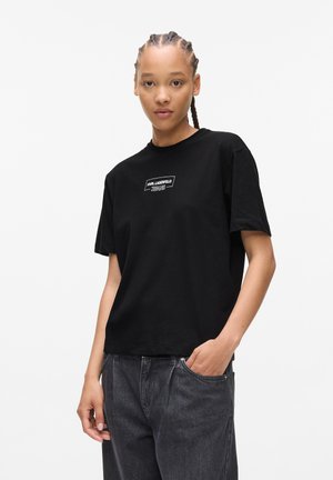 Black cotton t-shirt with a crew neckline, featuring a white logo on a rectangular patch. The fit is relaxed, paired with dark denim trousers.