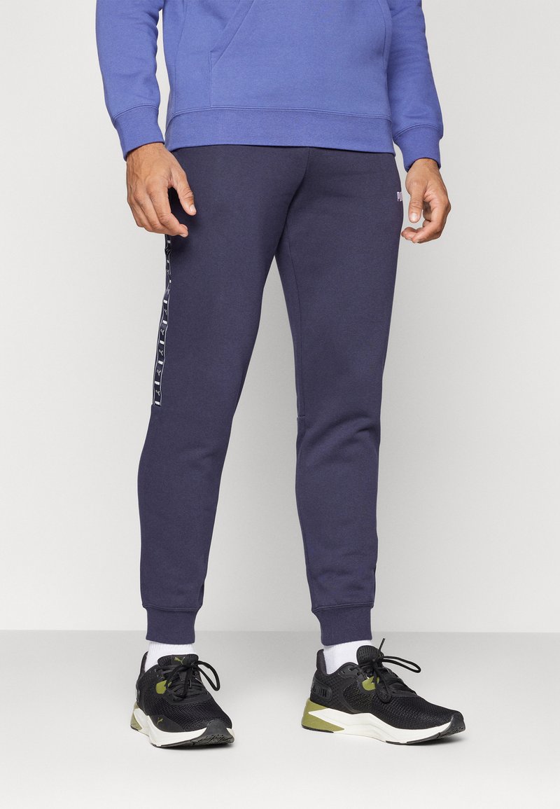 Marineblaue Jogginghosen mit schmaler Passform, seitlichen Streifen, gerippten Bündchen und weicher Textur. Getragen mit schwarzen Sportschuhen und weißen Socken.