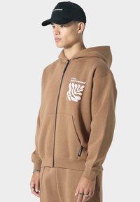 Brauner Reißverschluss-Hoodie aus weichem Material, mit einem weißen Blattgrafik und Logo auf der Brust, Fronttasche und gerippten Bündchen.
