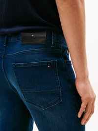 Jeans azul-escuro feitos de denim, com um patch de couro na parte de trás da cintura e um pequeno detalhe de logotipo vermelho, branco e azul no bolso.