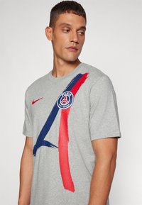 Sivi majica s kratkim rukavima koja sadrži višebojni dizajn poteza kistom u plavoj i crvenoj boji, s logom Paris Saint-Germaina na prednjem dijelu.