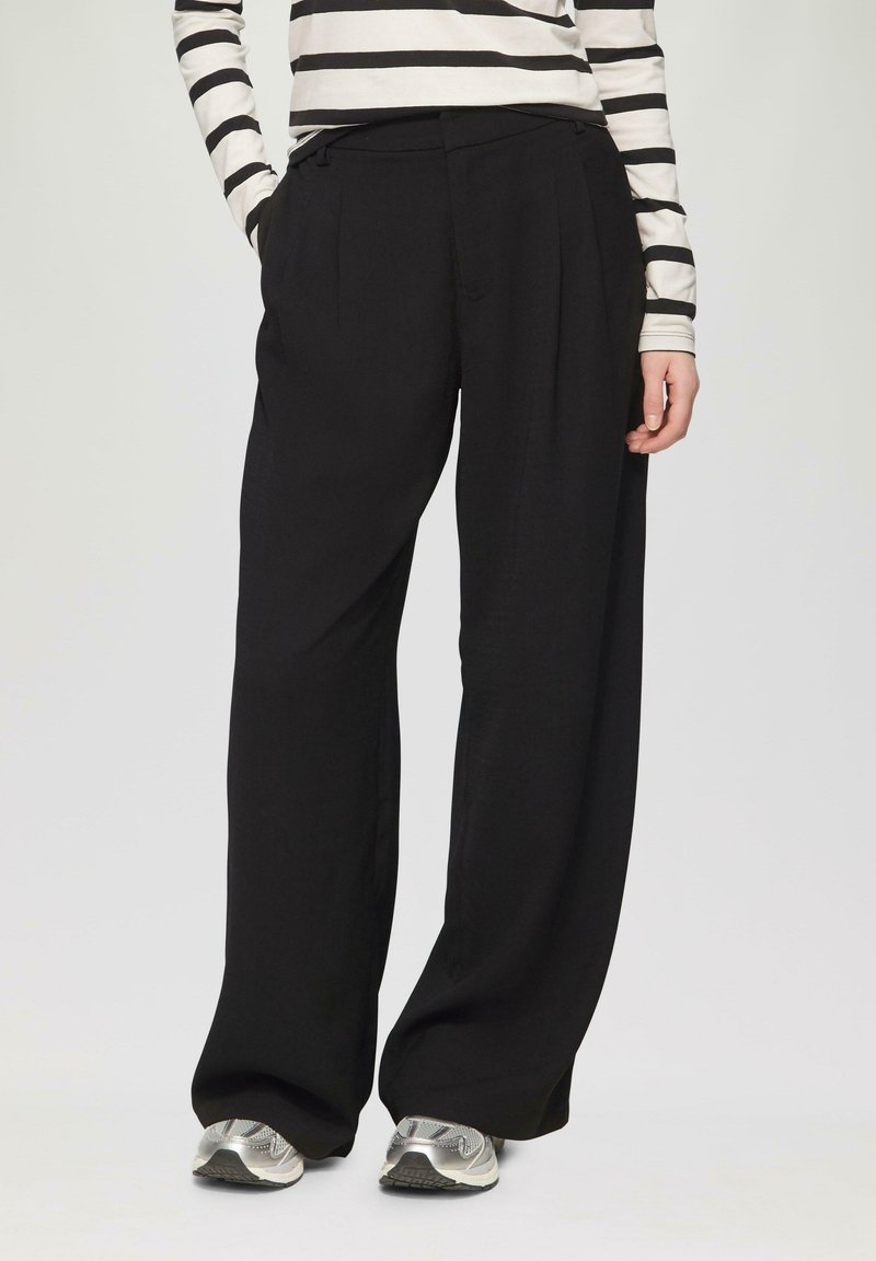 Schwarze weite Hose mit hoher Taille, Falten und einer glatten Textur, kombiniert mit silbernen Sportschuhen und einem gestreiften Langarmoberteil.