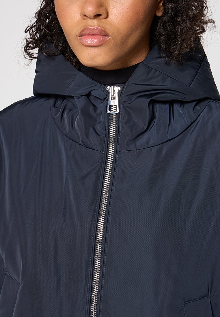 Marineblauwe pufferjack met een hoge kraag, ritsluiting en geplooide capuchon. Gladde, glanzende textuur en zijzakken. Zilveren accenten.