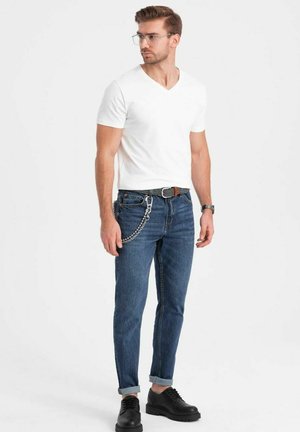 Weißes V-Ausschnitt-T-Shirt, blaue Jeans mit hochgerollten Säumen, schwarzer Gürtel mit Kettendetails und schwarze Schnürschuhe.