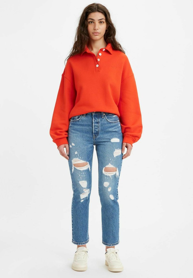Felpa oversized arancione con colletto e bottoni bianchi, abbinata a jeans blu strappati. Sneakers color crema completano il look.
