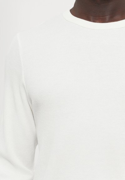 Chemise blanche à manches longues avec une texture et un motif côtelé, accompagnée d'un col contrastant. Le tissu semble léger et respirant.