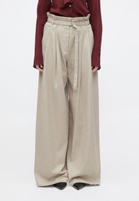 Pantalon beige taille haute à jambes larges avec ceinture en tissu, porté avec un chemisier bordeaux à manches longues et des chaussures noires pointues.