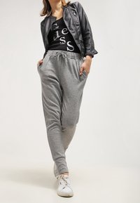 Kobieta idąca z rękami w kieszeniach, ubrana w czarną skórzaną kurtkę, czarny t-shirt z grafiką, szare dresy i białe trampki na jednolitym tle.