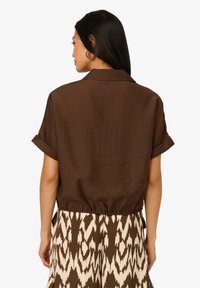 Chemise marron à manches courtes avec col et taille cintrée. Le tissu a une finition légèrement texturée. Portée avec une jupe fluide à motif.