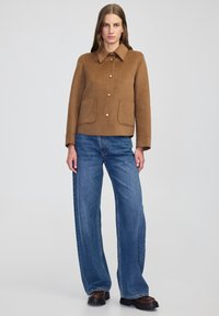 Brązowa kurtka z mieszanki wełny z kołnierzykiem, z zapięciem na guziki z przodu oraz dwoma przednimi kieszeniami, zestawiona z niebieskimi spodniami o szerokich nogawkach i czarnym obuwiem.