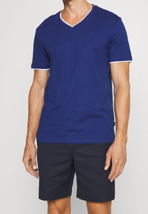 Homme portant un T-shirt bleu col en V à manches courtes avec bordure blanche et un short bleu marine foncé sur un fond uni.