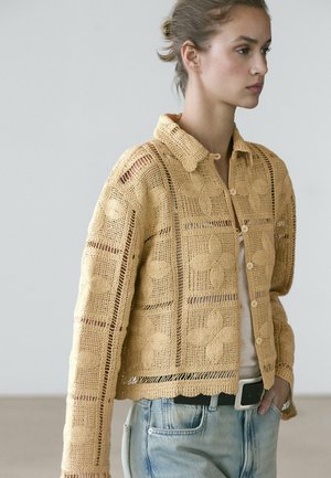 Vrouw draagt een beige gebreid vest met knopen en bloemmotieven over een wit shirt, gecombineerd met lichtblauwe jeans en een zwarte riem.