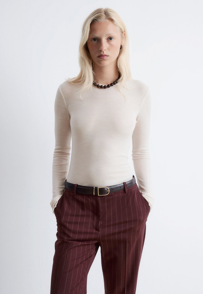 Cremfarbenes Langarmoberteil aus leichtem Stoff, kombiniert mit dunkelburgunderfarbenen Nadelstreifenhosen und einem schwarzen Gürtel, ergänzt durch eine runde Halskette.