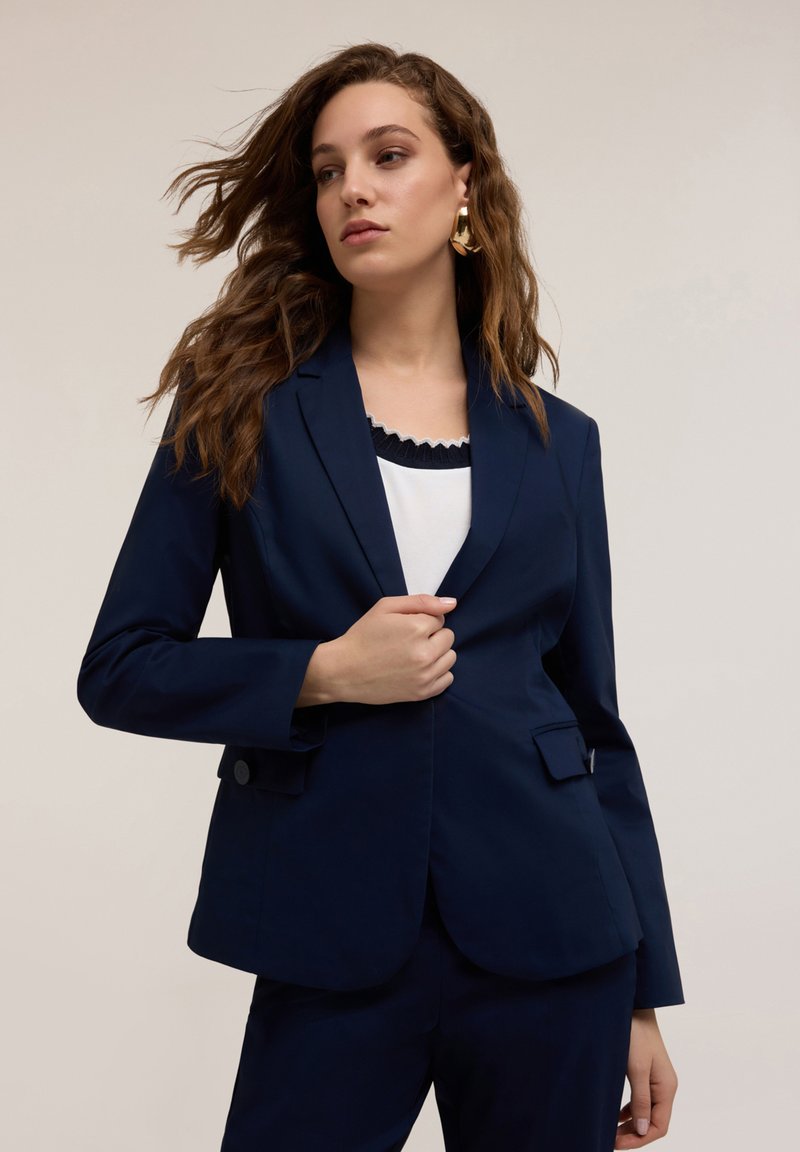 Blazer navy con un corte entallado, solapas de pico y dos bolsillos frontales. Llevado sobre una blusa blanca con un cuello fruncido, que presenta mangas largas.