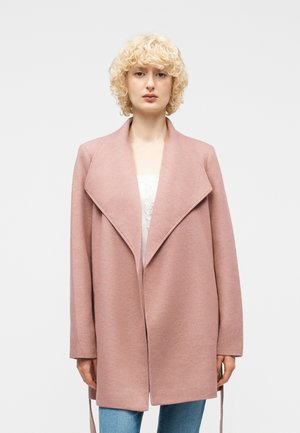 Persona con capelli biondi e ricci che indossa un cappotto largo color rosa polvere con ampi risvolti sopra jeans blu, in piedi davanti a uno sfondo chiaro e uniforme.