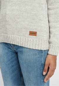 Lichtgrijze gebreide trui met geribde zoom en mouwen. Bevat een leren label met "oxmo" branding aan de voorkant. Draagt over blauwe jeans.