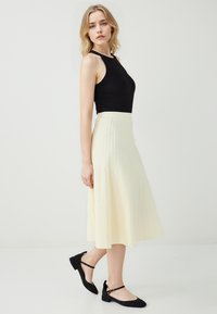 Top noir sans manches à col montant, associé à une jupe plissée midi de couleur jaune clair. Des ballerines noires avec des brides de cheville viennent compléter le look.