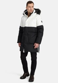 Padded parka ar baltu augšējo daļu un melnu apakšējo daļu, ar kažokādas oderētu kapuci, priekšējiem kabatām un rāvējslēdzēja akcentiem.
