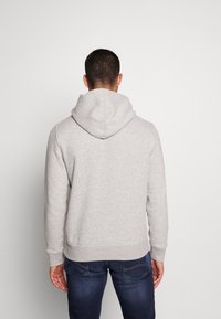 Uomo di spalle che indossa un hoodie grigio chiaro e jeans blu scuro su uno sfondo chiaro e semplice.