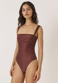 Brązowy, żebrowany jednoczęściowy strój kąpielowy z kwadratowym dekoltem i szerokimi ramiączkami. Posiada niskie wycięcia na biodrach i gładką teksturę.