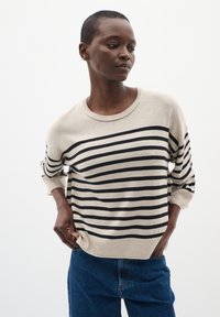 Maglione beige con righe orizzontali nere, scollatura rotonda e maniche a tre quarti. Indossato con jeans blu. Tessuto morbido.
