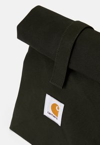 Olivově zelená látková taška s rolovaným vrškem a suchým zipem. Obsahuje bílou etiketu s oranžovým logem. Texturovaný plátěný materiál.