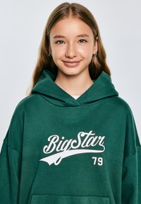 Zeleni oversized dukserica s prednjim džepom, s bijelim natpisom "Big Star 79". Mekana tekstura, kapuljača s vezicom i opušteni kroj.